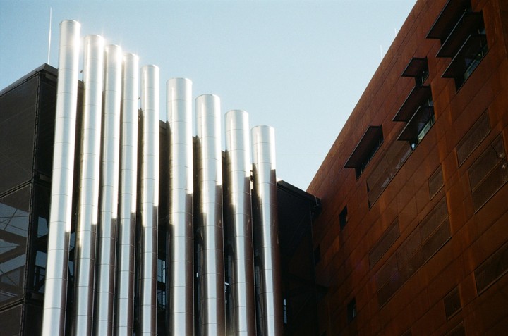 A modern building with shiny silver pipes on the left and a rusty orange facade on the right stands against a clear sky.