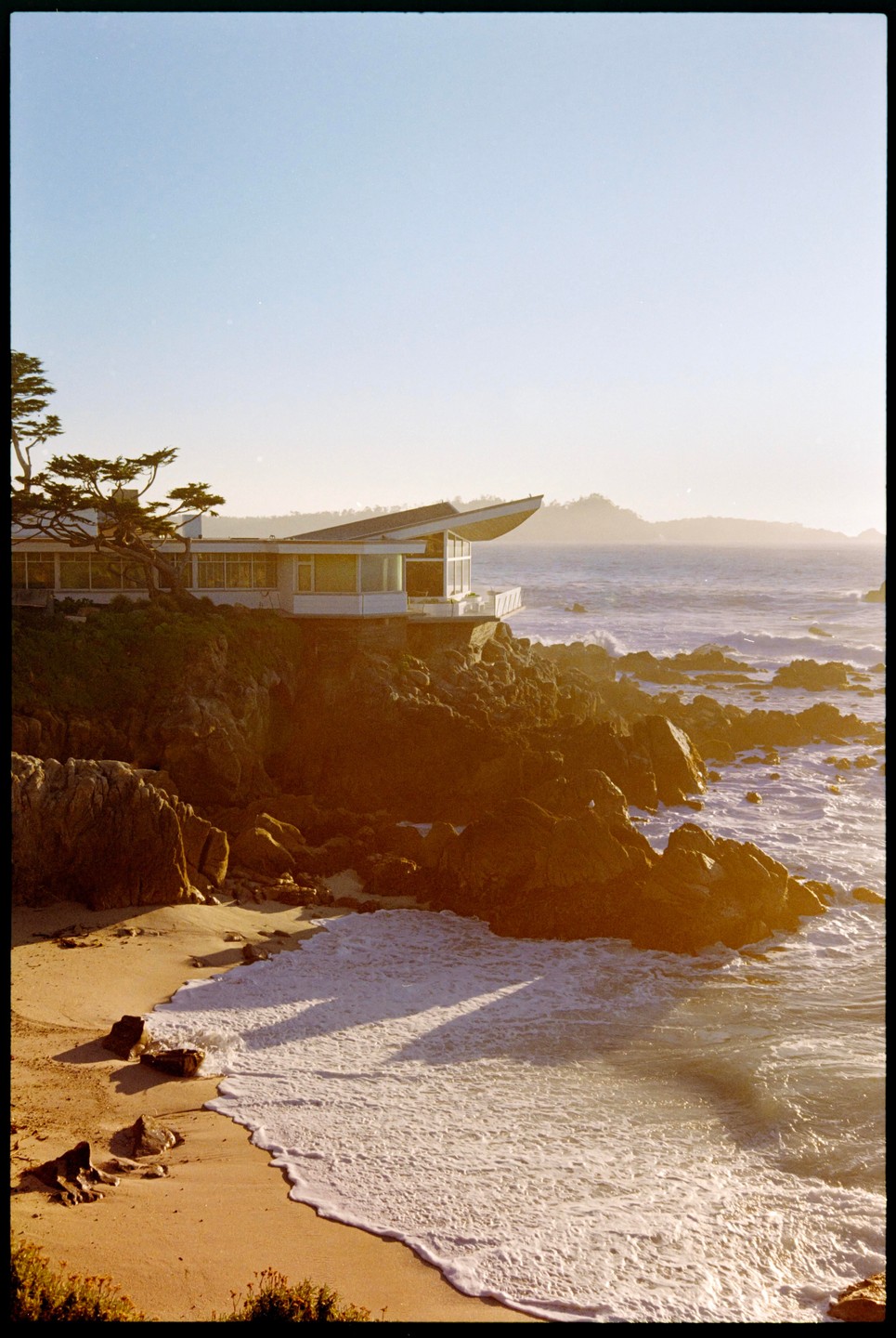 A modern house sits on a rocky cliff overlooking a sandy beach with waves crashing on the shore.