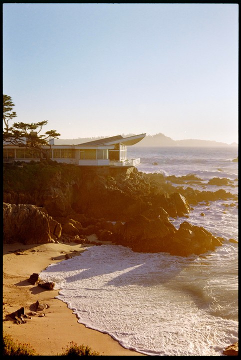 A modern house sits on a rocky cliff overlooking a sandy beach with waves crashing on the shore.