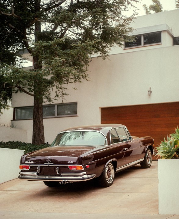 A brown vintage car is parked in a driveway next to a modern white building with a wooden garage door.