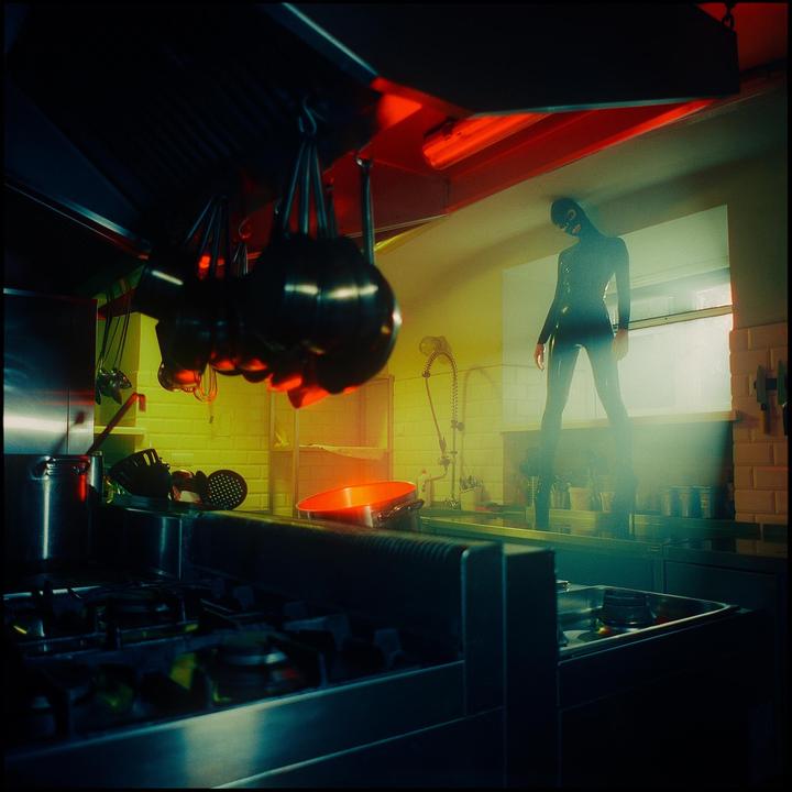 A person in a black latex suit and mask stands in a kitchen with red and yellow lighting.