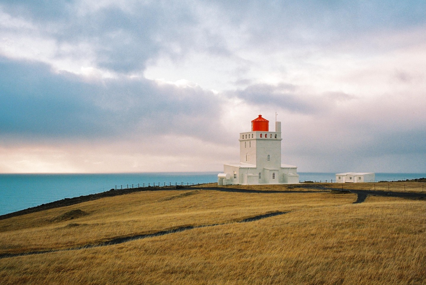 A white lighthouse with a red top stands on a grassy hill overlooking the ocean under a cloudy sky.