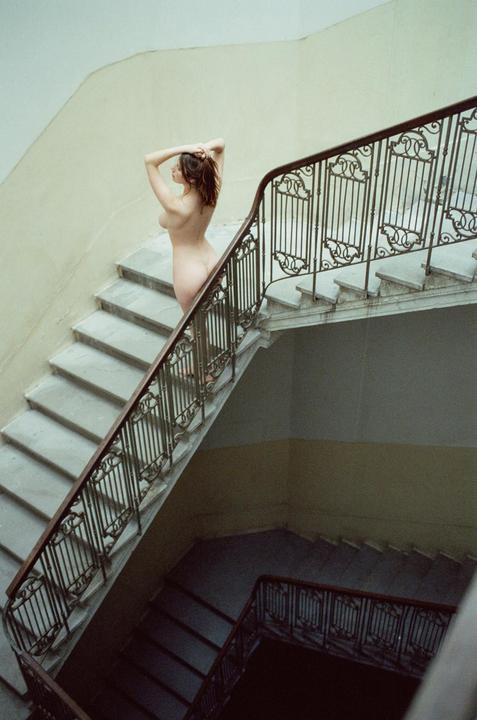 A nude woman stands on a marble staircase with an ornate railing, looking away from the camera.