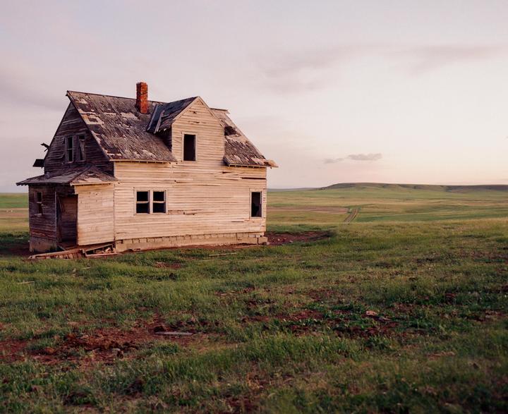 An abandoned house with a red brick chimney stands in a grassy field under a pale sky.
