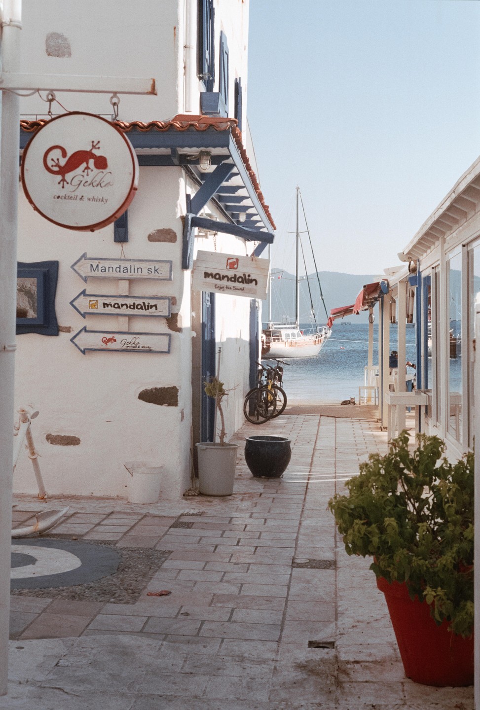 A narrow alleyway between white buildings leads to a sailboat docked by the sea.