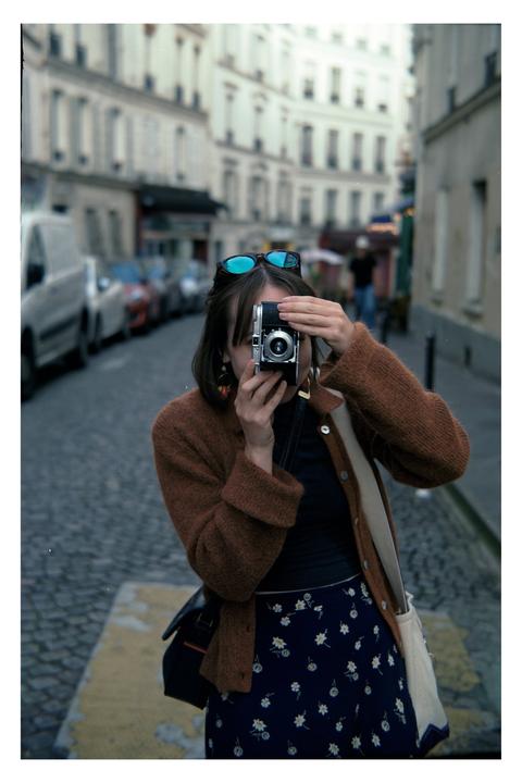 A woman holds a camera up to her face on a cobblestone street with buildings and parked cars in the background.