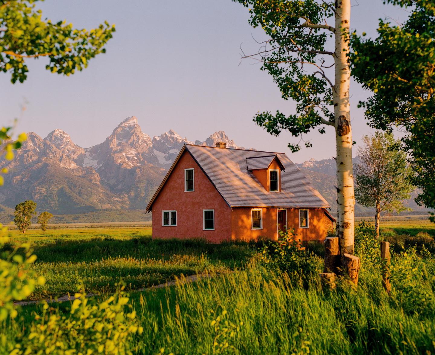 A red house stands in a green field with snow-capped mountains in the background under a clear sky.
