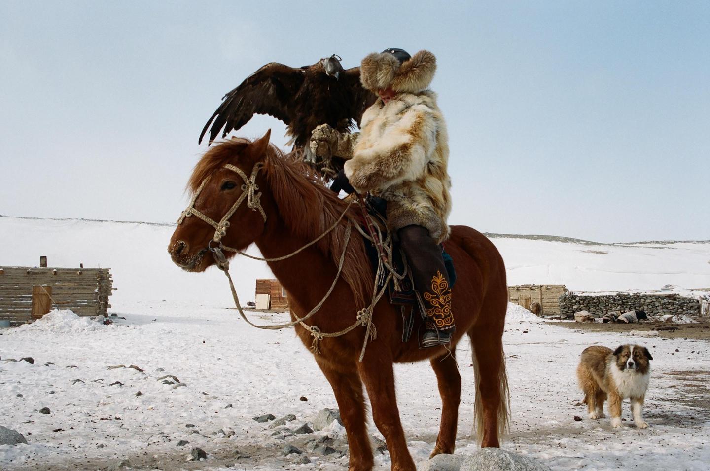 A person in a fur coat and hat rides a horse with an eagle on their arm in a snowy landscape, with a dog standing nearby.