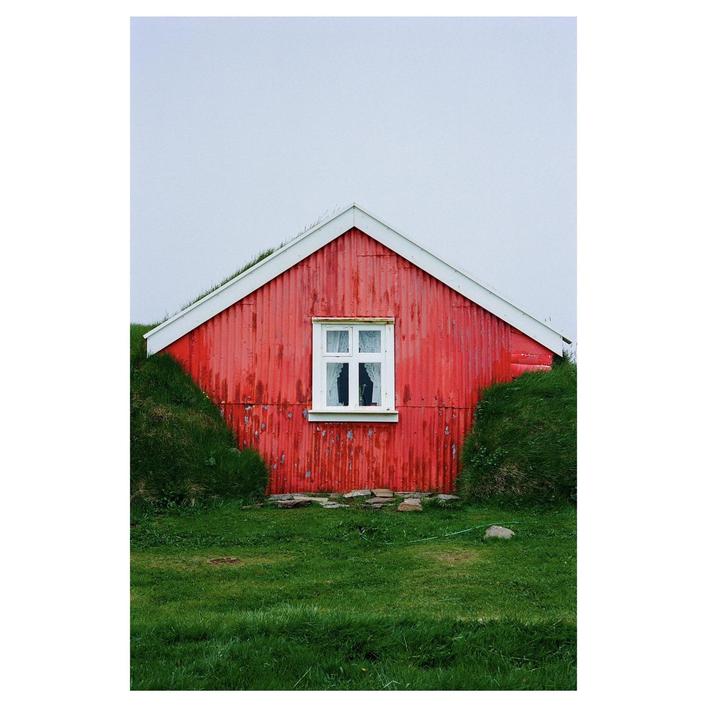 A red house with a white window is surrounded by green grass under a light sky.