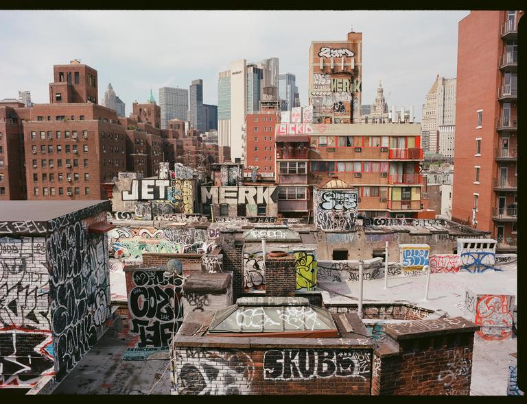 A cityscape shows many buildings with graffiti on their rooftops and walls, with taller buildings in the background.