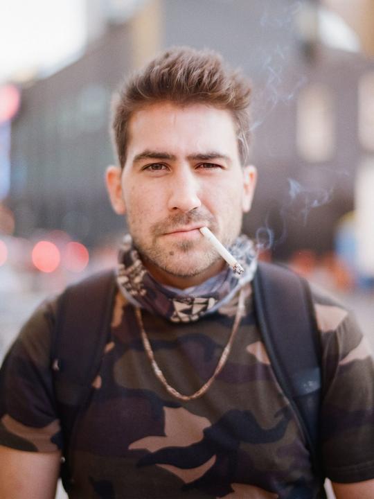 A man with a cigarette in his mouth looks directly at the viewer, with smoke rising from the cigarette.