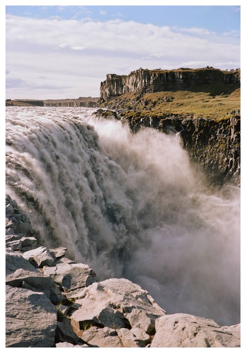 A powerful waterfall cascades over a rocky cliff into a misty gorge, with a flat, grassy landscape and a cloudy sky above.