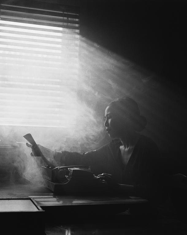 A woman sits at a desk with a typewriter, illuminated by light streaming through a window blind.