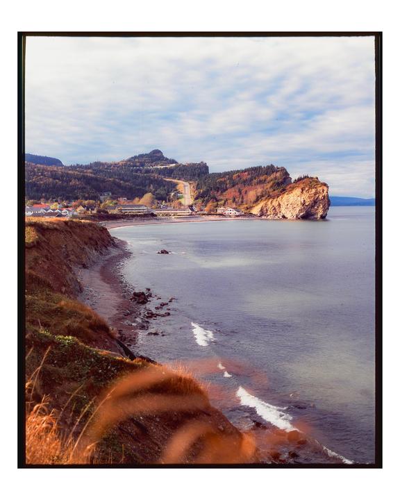A coastal town with a road winding through hills and a large rock formation extending into the water is visible under a cloudy sky.