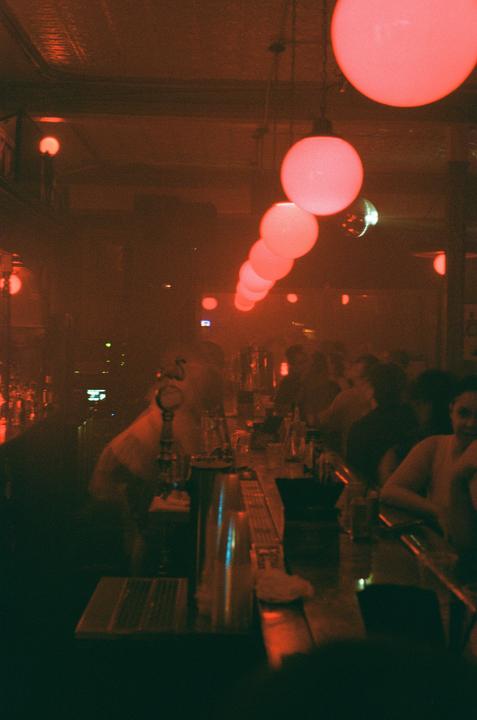 A long bar is lit by a row of red globe lights hanging from the ceiling, with people sitting and standing along its length.
