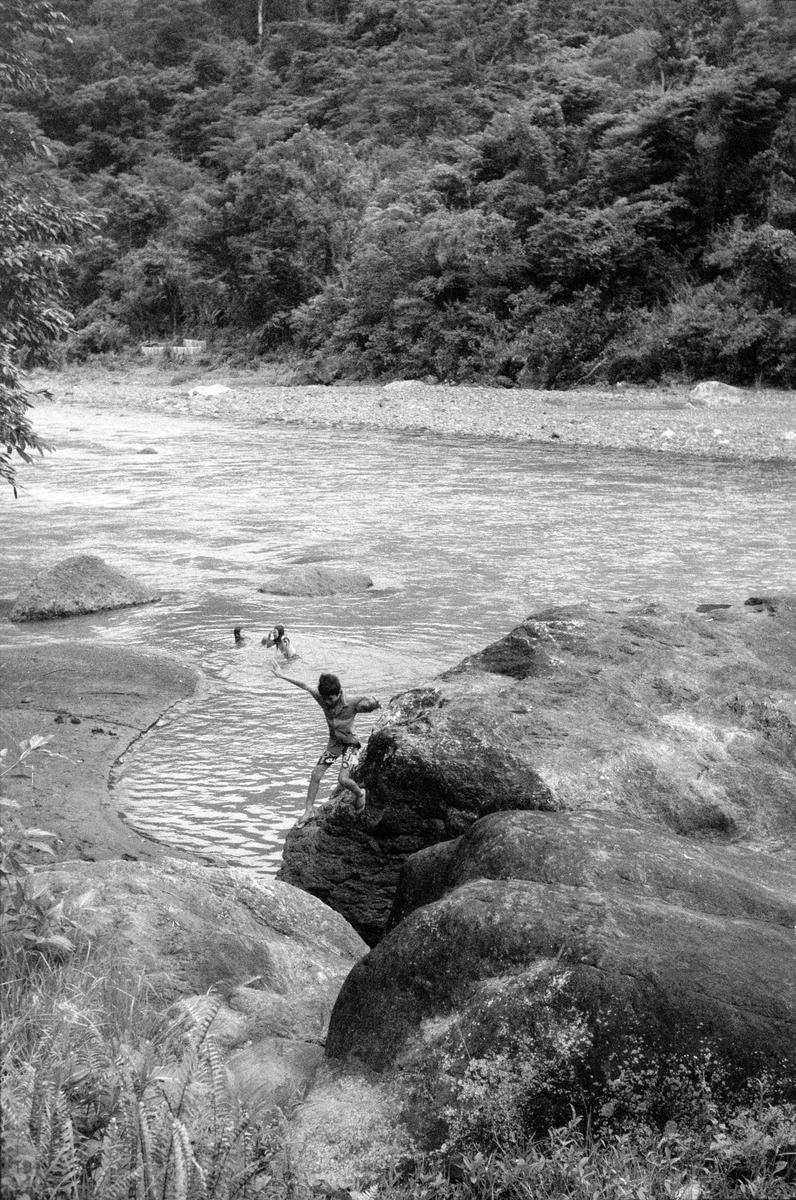 A child stands on a rock with arms outstretched, looking towards other children swimming in a river, with a forested hill in the background.