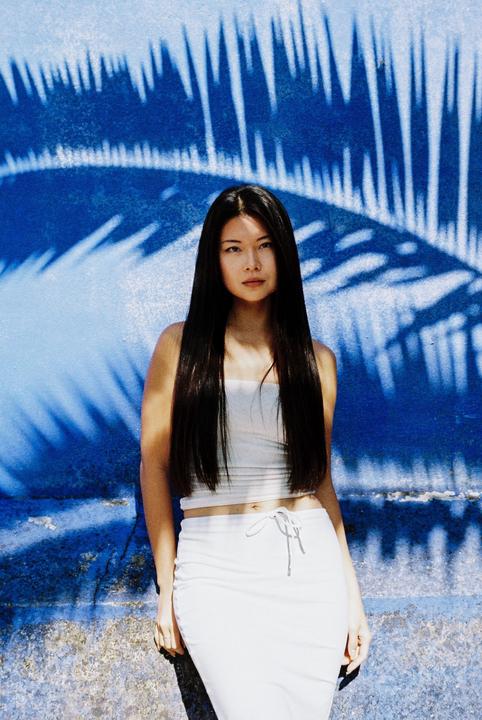 A woman with long dark hair stands in front of a blue wall with palm tree shadows.