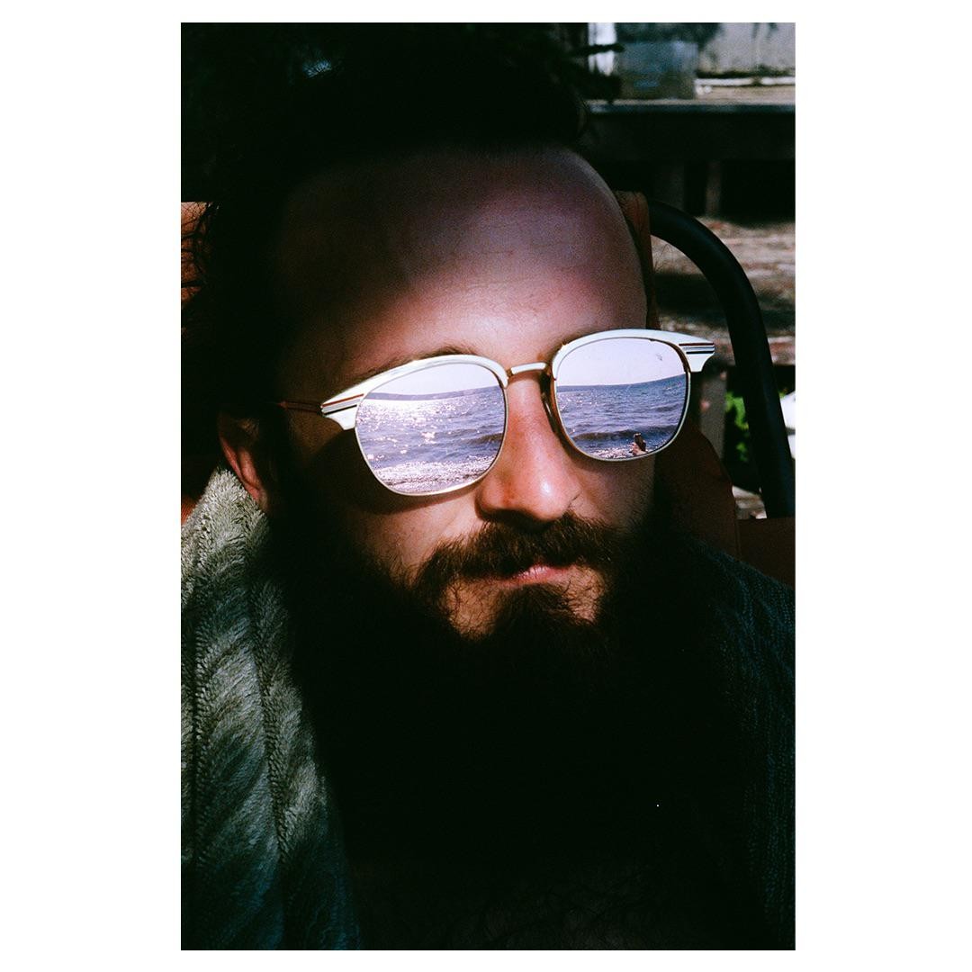A man with a beard and sunglasses has a reflection of the ocean in his lenses.