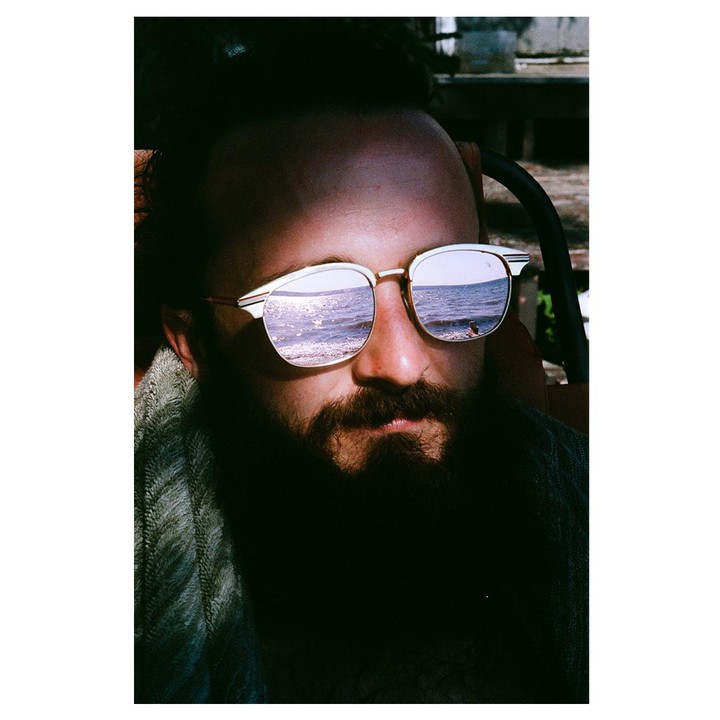 A man with a beard and sunglasses has a reflection of the ocean in his lenses.