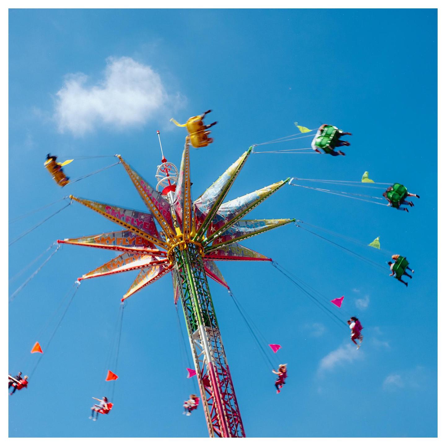 People ride a colorful swing ride against a bright blue sky with a single cloud.
