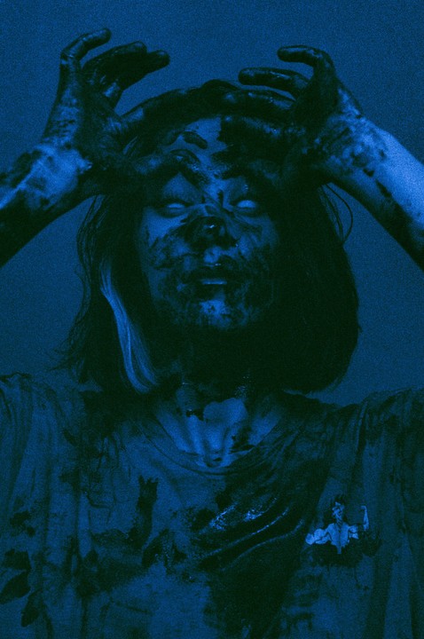 A person with long hair and a dirty face holds their hands up to their head, all in a blue monochrome.