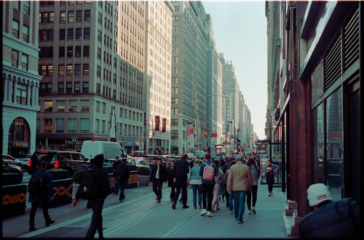 A busy street scene shows pedestrians walking on a sidewalk next to tall buildings and parked cars.