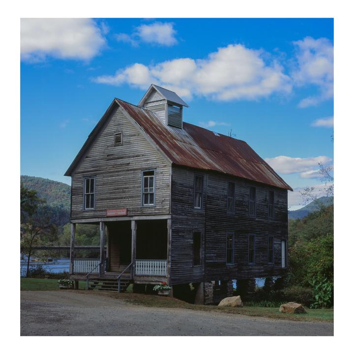 An old wooden meeting hall with a rusty tin roof stands beside a river with mountains in the background under a blue sky with white clouds.