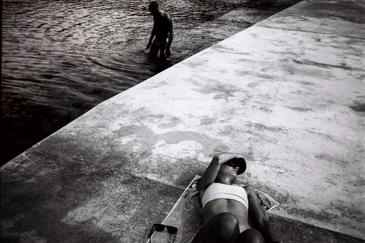 A woman sunbathes on a concrete surface next to water where a man is standing.