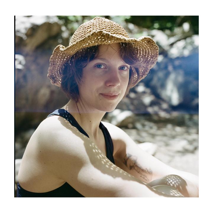 A woman wearing a straw hat sits outdoors with dappled sunlight on her arms.