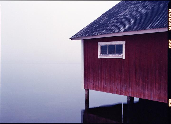 A red boathouse with a dark roof stands over calm water under a hazy sky.