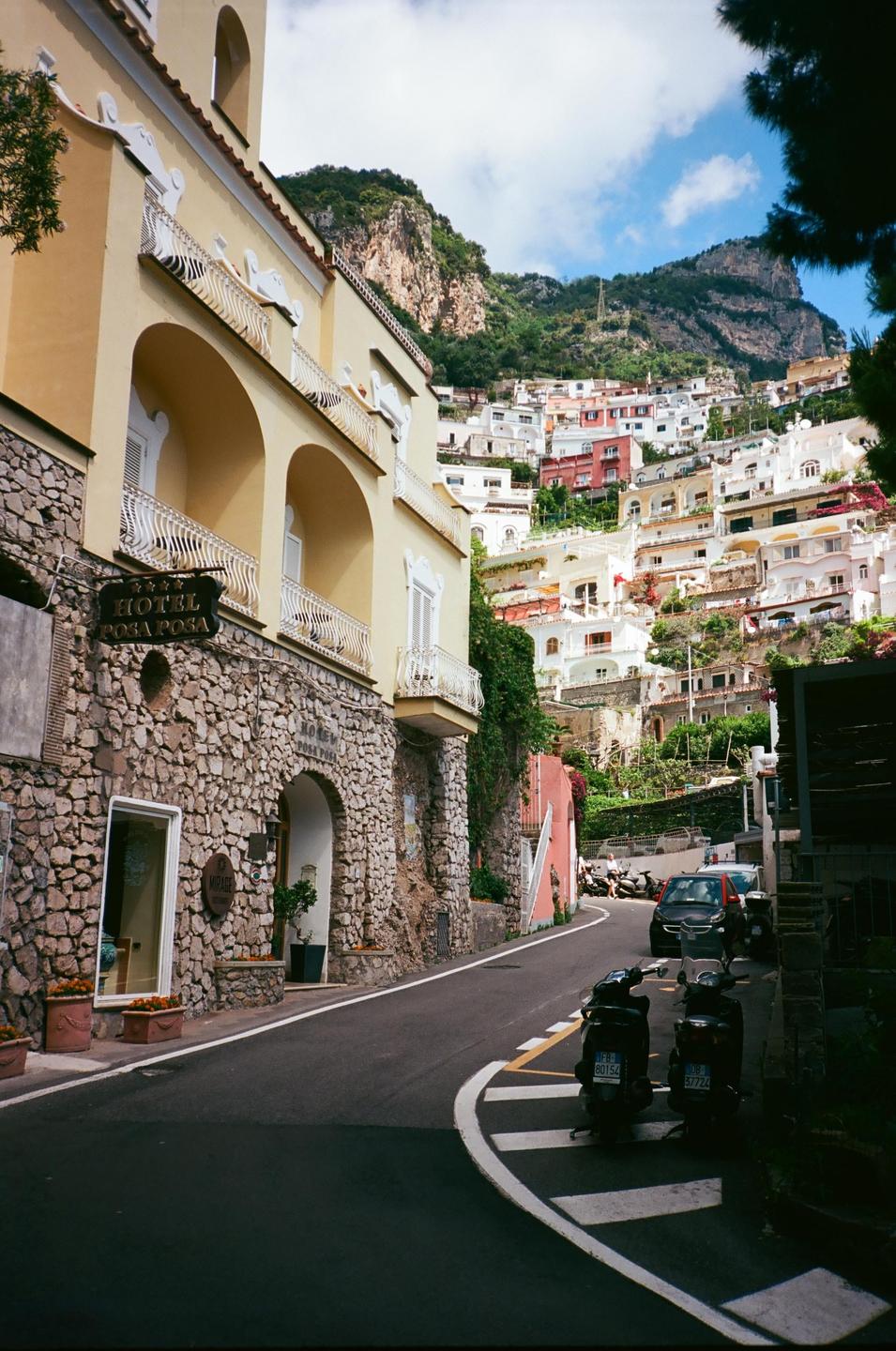 A narrow street winds past a stone building with a hotel sign, leading up to a hillside town with many colorful houses.