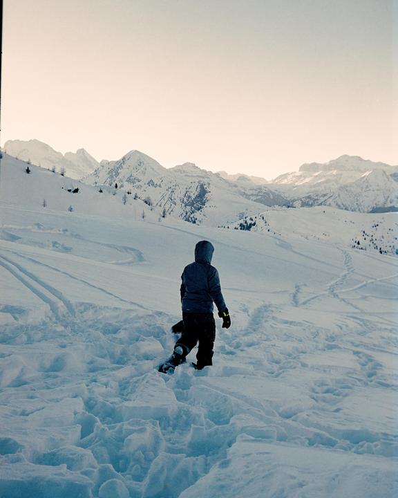 A person in a hooded jacket walks through deep snow with mountains in the background.