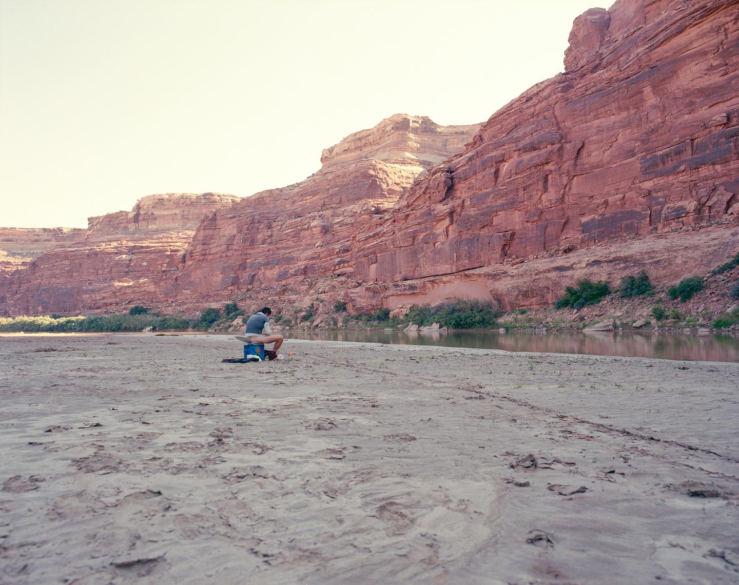 A man sits on a blue cooler on a sandy riverbank with large red rock formations in the background.