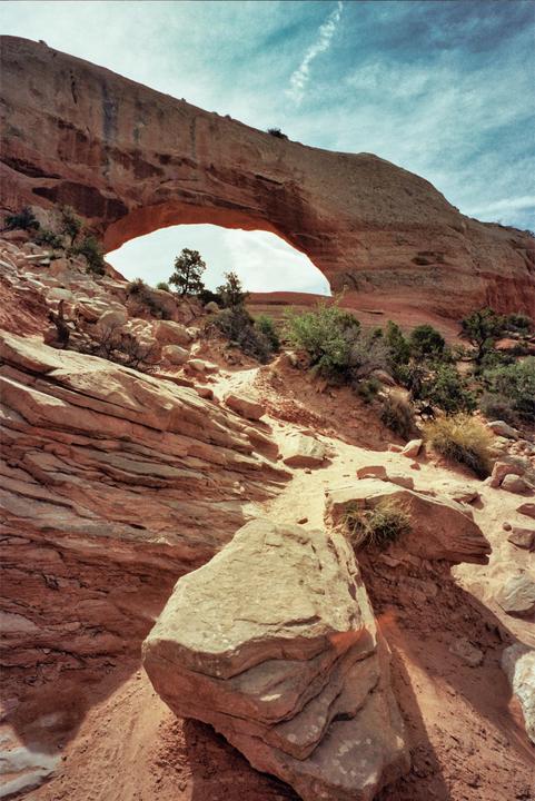 A natural arch is visible through a rocky landscape with sparse vegetation under a cloudy sky.
