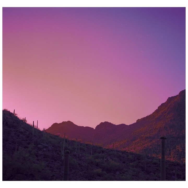 A desert landscape with mountains and cacti is illuminated by the vibrant colors of a sunset sky.