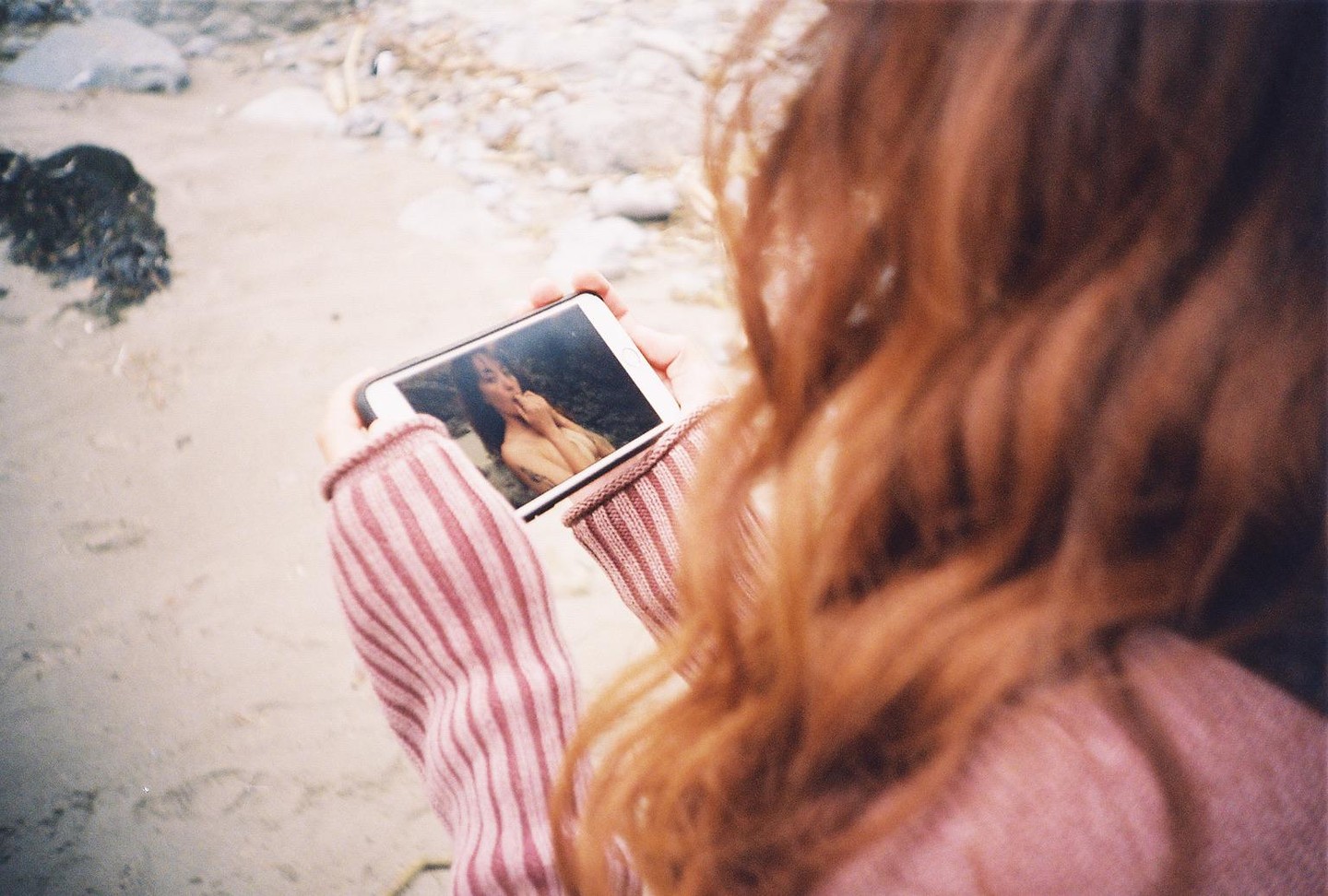 A woman with long red hair holds a smartphone displaying a photo of a woman on a beach.