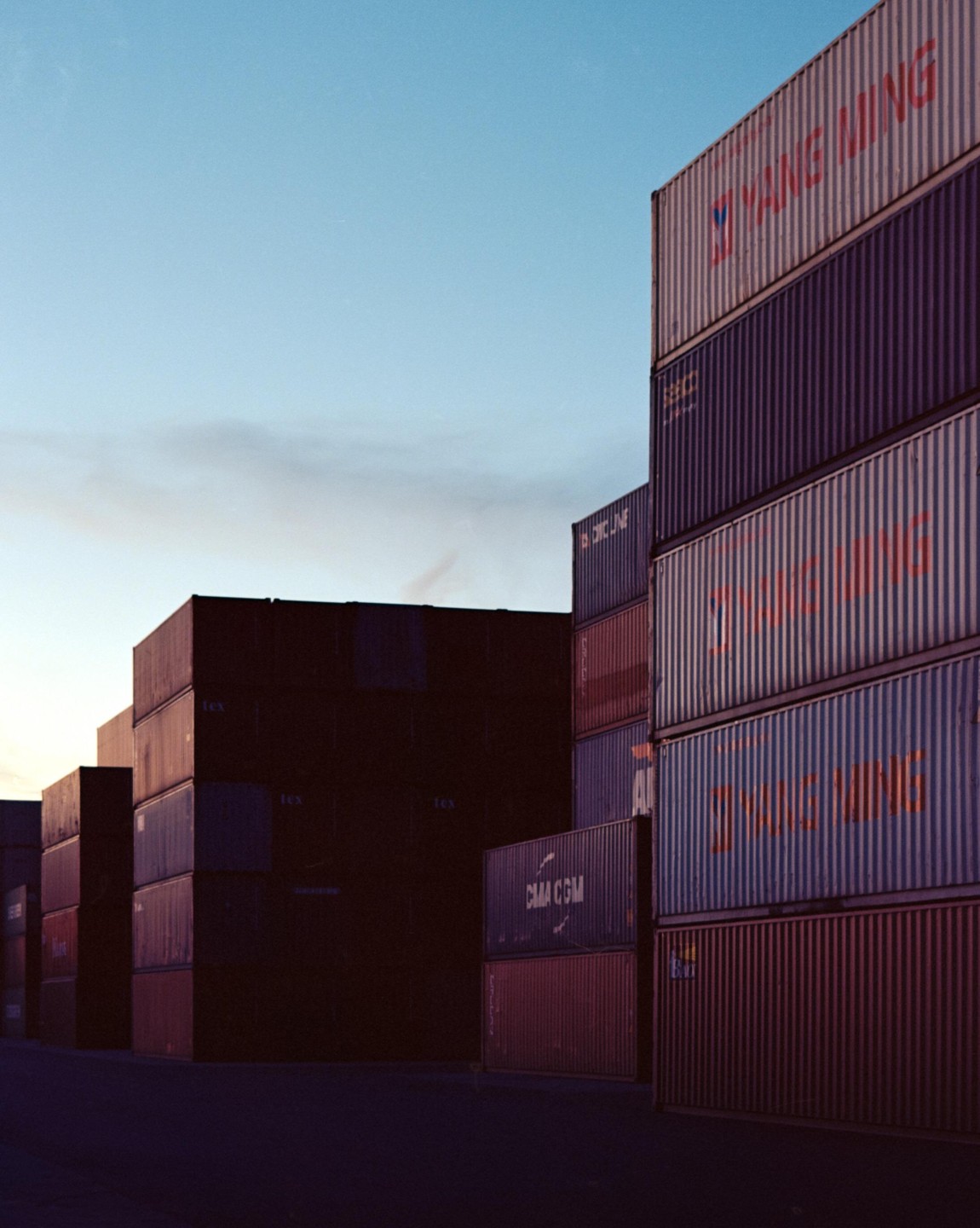 Tall stacks of shipping containers are silhouetted against a clear sky at dusk.