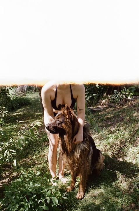 A woman in a bikini top stands in a garden, holding a large dog.