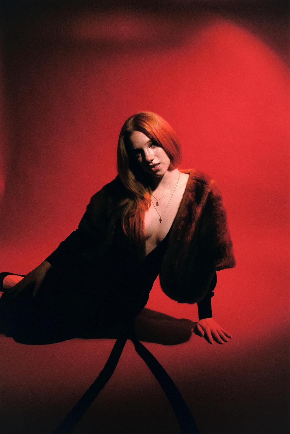 A woman with long hair and a fur stole is sitting on the floor against a red background.