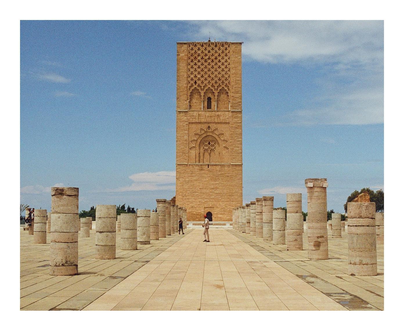 A tall, ornate tower stands behind rows of stone pillars on a paved plaza under a blue sky.
