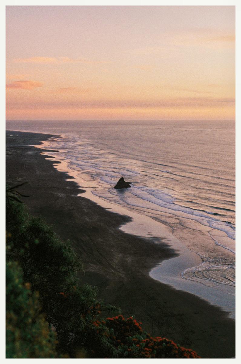 A high-angle landscape view shows a beach and ocean at sunset with a single rock formation in the water.