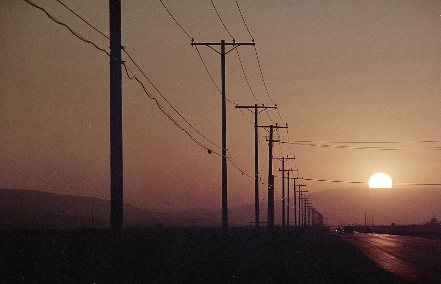 A row of utility poles and power lines stretches along a road with a large sun setting in the distance.