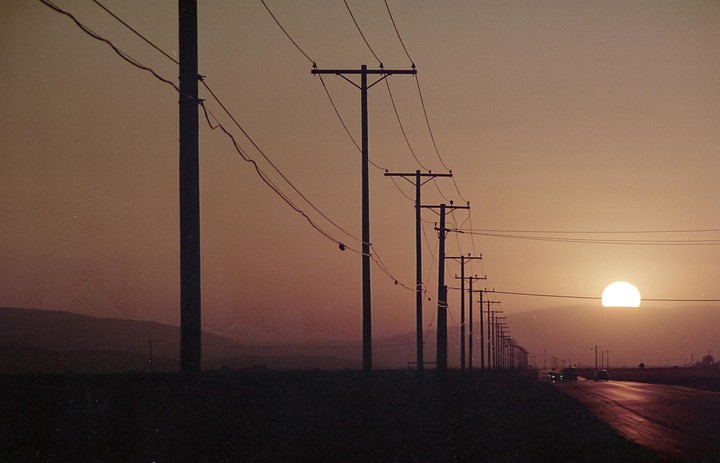 A row of utility poles and power lines stretches along a road with a large sun setting in the distance.