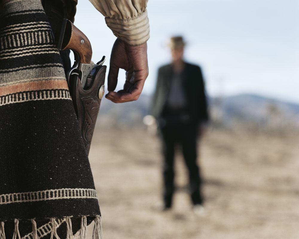 A man with a revolver in a holster stands in a desert landscape, facing another man in the distance.