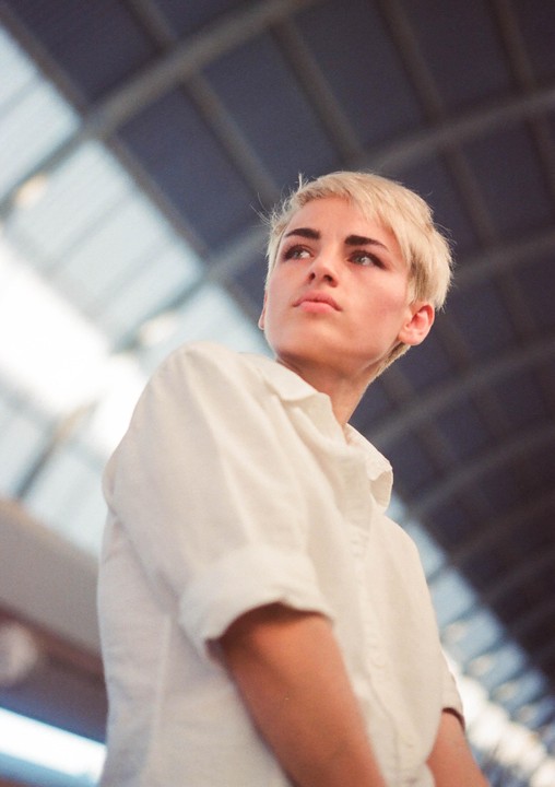 A woman with short blonde hair and a white shirt looks up and to the right, with a blurred railway station roof in the background.