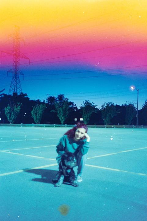 A woman and a dog are crouching on a parking lot at dusk, with a colorful light leak across the top of the image.