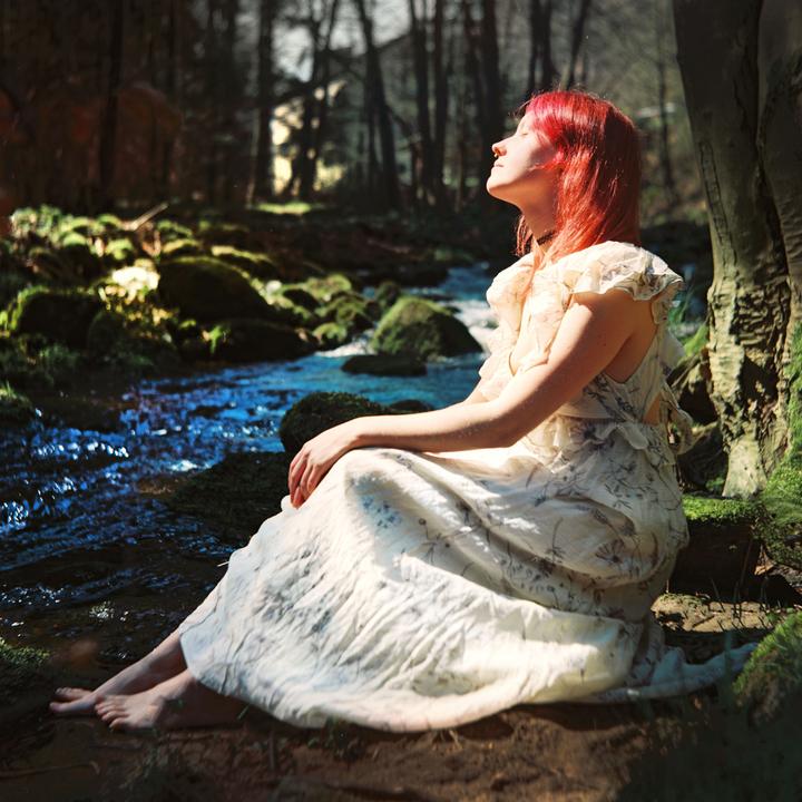 A woman with red hair sits by a stream in a forest, looking up with her eyes closed.