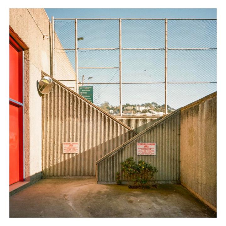 A red door and a convex mirror are next to a concrete staircase with a chain-link fence above it.