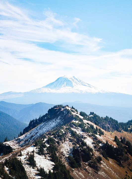 A snow-capped mountain rises in the distance behind a ridge covered with trees and patches of snow.