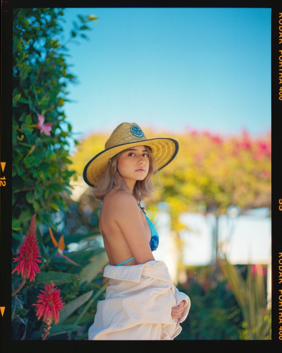 A woman wearing a straw hat and a blue bikini top stands among green foliage and red flowers.